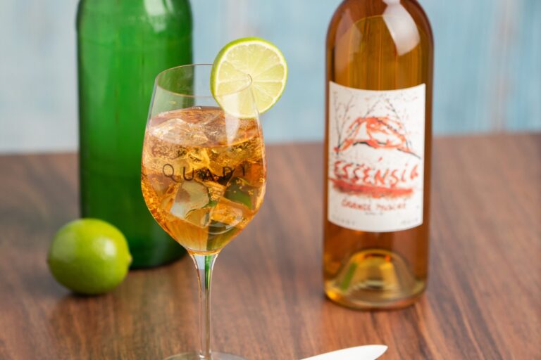 Essensia Spritzer wine cocktail in a wine glass on a table next to a lime, bottle of Essensia Orange Muscat Dessert wine and bottle of club soda.