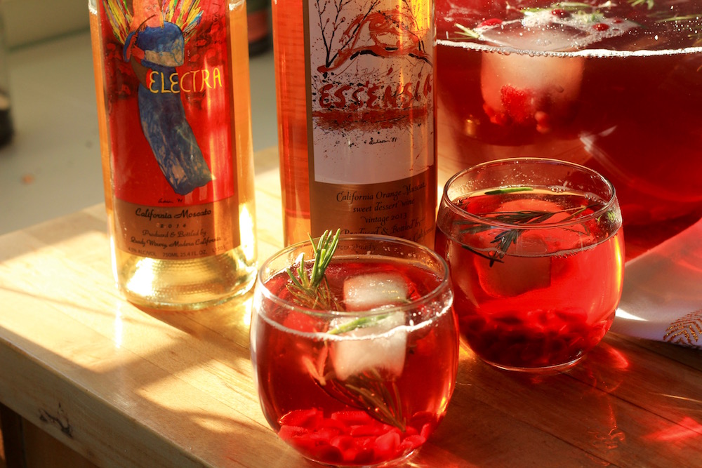 Jingle Juice cocktail in two glasses filled with ice next to a punch bowl, bottle of Essensia Orange Muscat sweet dessert wine and Electra Moscato wine.