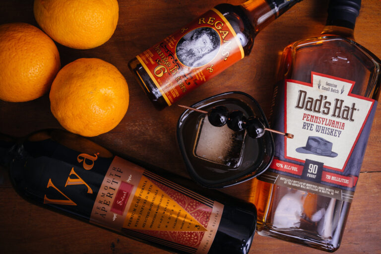 Manhattan cocktail in a cocktail glass filled with a an ice cube and maraschino cherry on a table next to a bottle of Vya Sweet Vermouth, Dad's Hat Rye Whiskey, Regan's Orange Bitters and whole oranges.