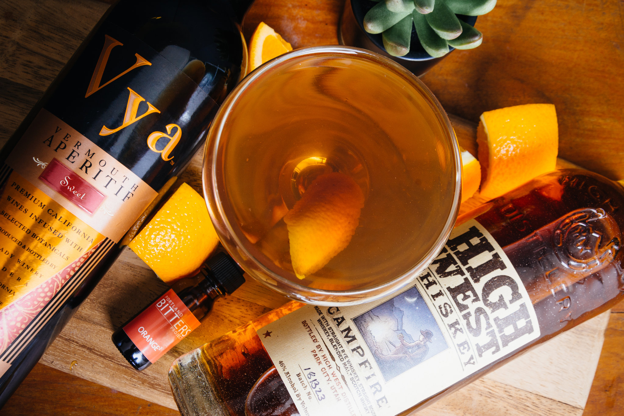 Friends and neighbors manhattan cocktail made with High West Campfire Whiskey and Vya Sweet Vermouth in a coupe glass with an orange peel garnish on a table next to a bottle of Vya Sweet Vermouth, High West Whiskey, Bitters and orange slices.