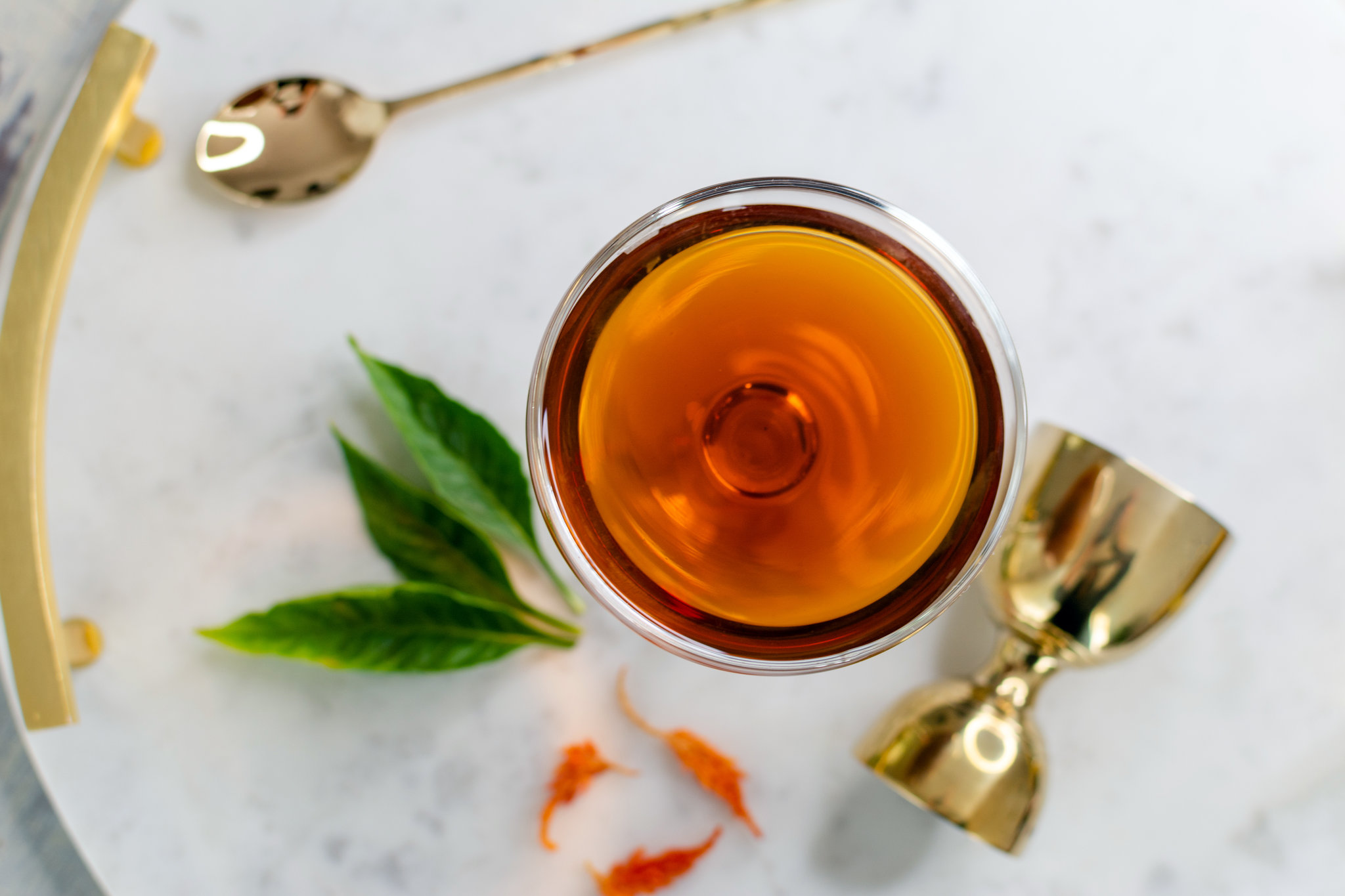 Maple Manhattan cocktail in a coupe glass next to stir spoon, jigger, and various herbs.