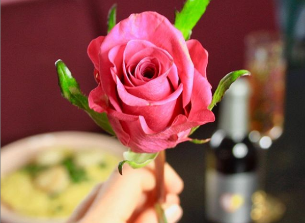 Hand holding rose with a bottle of Elysium Black Muscat dessert wine and meal in back ground.
