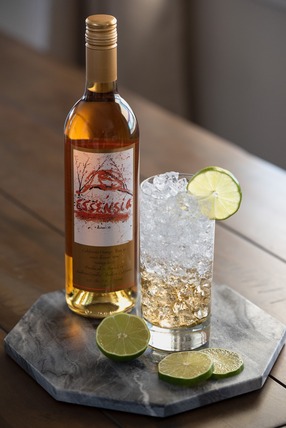 Bottle of Essensia Orange Muscat Dessert Wine on a table next to the Essensia Hard Seltzer cocktail with limes