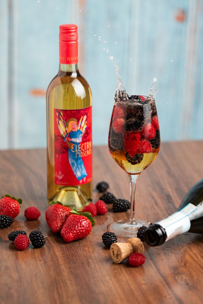 A wine glass filled with Electra Moscato, prosecco and fresh fruit sitting on a table with a bottle of Electra Moscato, some fresh fruit, and an empty bottle of prosecco next to it.