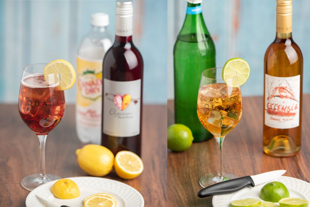 Elysium Spritzer wine cocktail in a wine glass on a table next to lemons, bottle of Elysium Black Muscat Dessert Wine and tonic water and Essensia Spritzer wine cocktail in a wine glass on a table next to a lime, bottle of Essensia Orange Muscat Dessert wine and bottle of club soda.