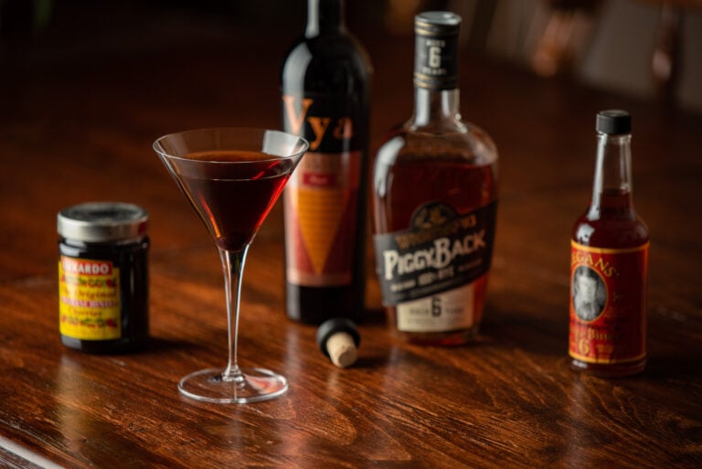 Manhattan Cocktail in a martini glass sitting on a table next to bottles of Vya Sweet Vermouth, Rye Whiskey, Bitters and Brandied Cherries.