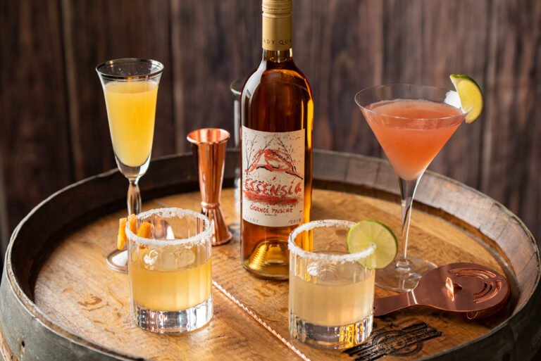 Bottle of Essensia Orange Muscat Dessert Wine sitting on a wine barrel surrounded by classic cocktails.