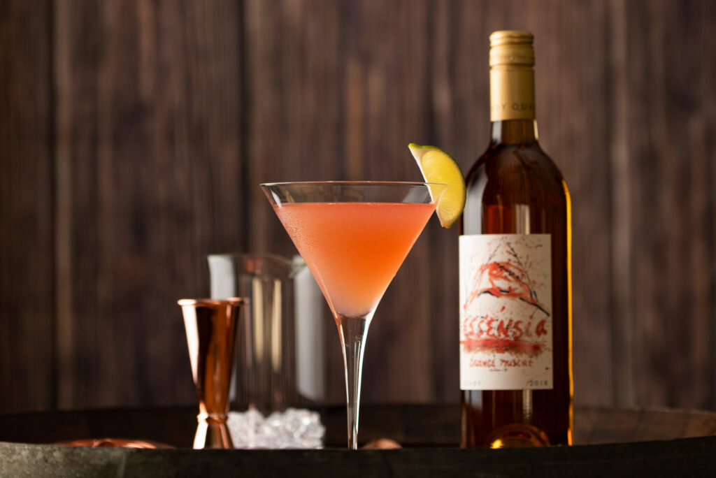 Essensia cosmopolitan cocktail in a martini glass next to barware and a bottle of Essensia Orange Muscat dessert wine by Quady Winery.