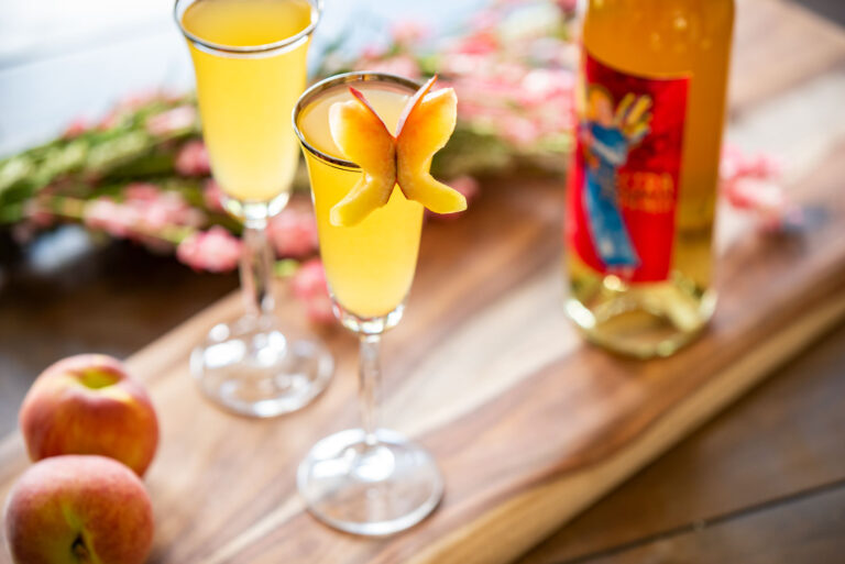 Electra Bellini wine cocktail in a champagne flute with a peach garnish next to a bottle of Electra Moscato wine.