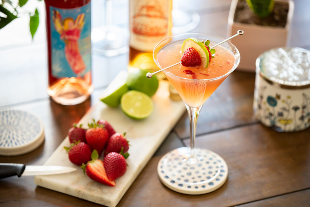 Pink Lady wine cocktail in a martini glass on a table next to ingredients and bottles of Electra Moscato Rosé and Essensia Orange Muscat Dessert Wine.