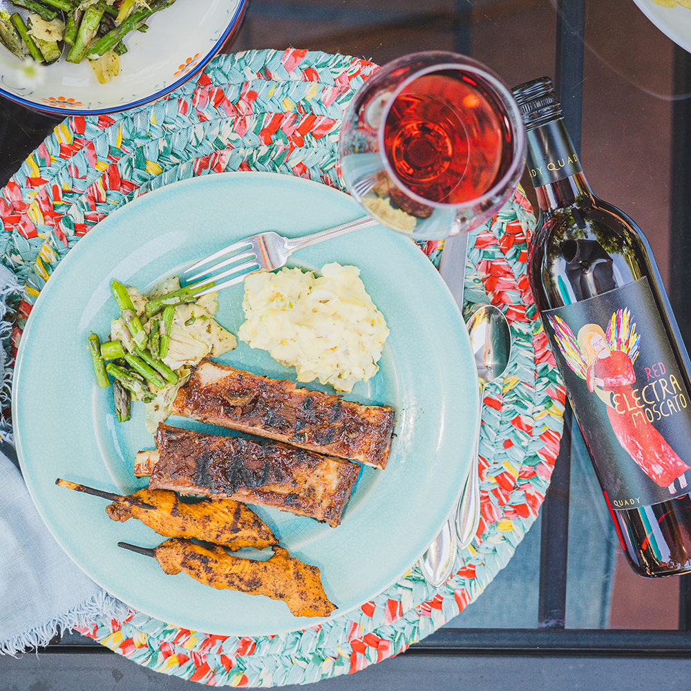 A glass of Red Electra Moscato with the bottle of wine and a plate of chicken, ribs, potato and veggie salads