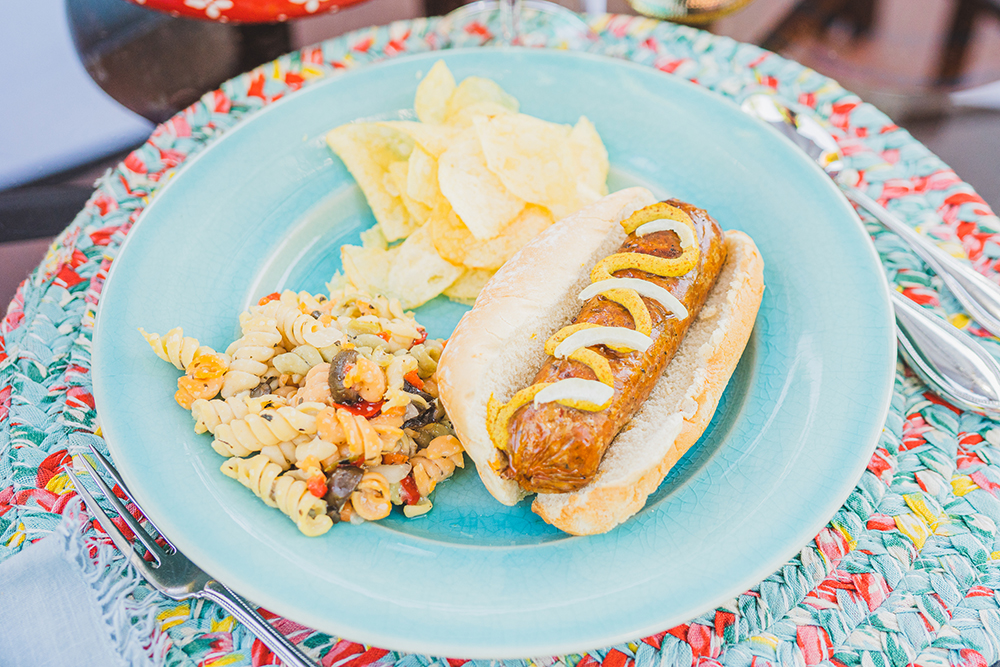 Brat in a bun on a plate with chips and pasta salad